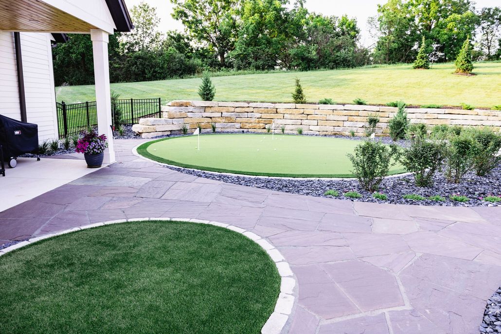 A backyard putting green with a stone patio, landscaping, and a tiered stone retaining wall in a grassy outdoor space.