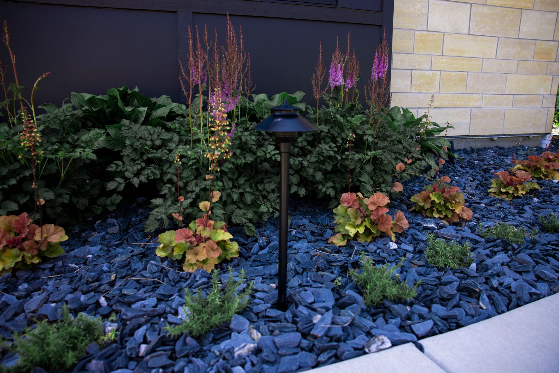 A garden bed with dark blue stones, astilbe flowers, coral bells, and a black path light against a stone wall.