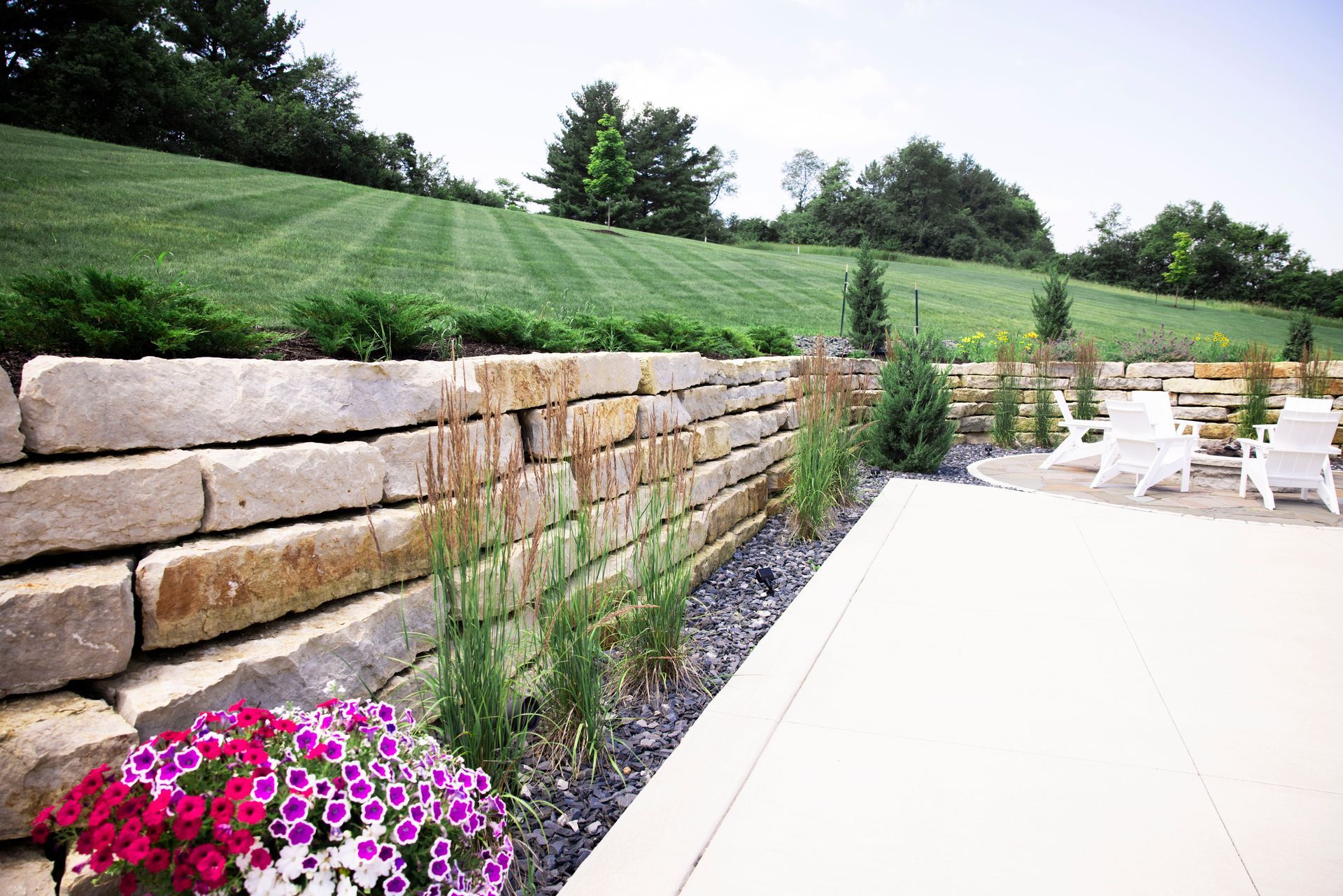 A stacked stone retaining wall borders a patio with white chairs, set against a backdrop of a mowed grassy hill.