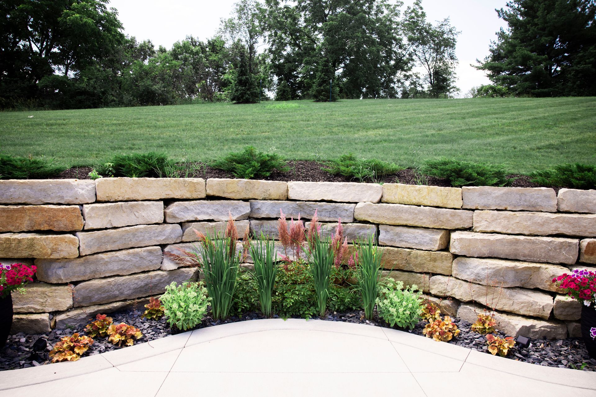 A curved stone retaining wall borders a patio, featuring a garden bed with ornamental grasses and flowers against a lawn.