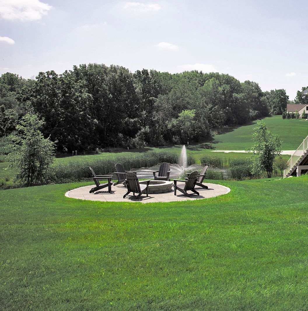 A patio area with Adirondack chairs and a fire pit, situated on a grassy lawn overlooking a pond with a fountain.