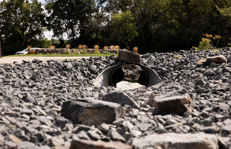 A culvert made of corrugated metal drains water through a bed of gravel under a road.