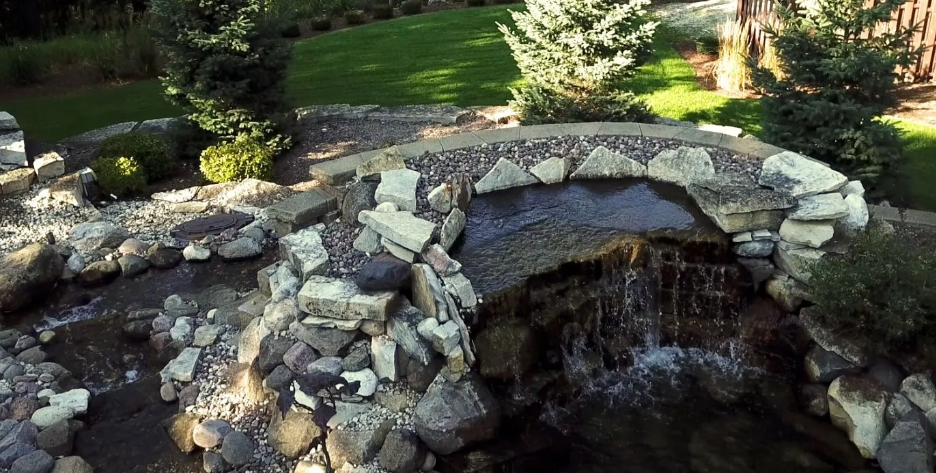 Stone bridge over a waterfall in a backyard, surrounded by rocks and greenery.