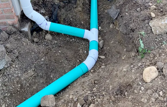 Green and white pipes in a trench, connected with elbows and a tee joint; near a brick wall.