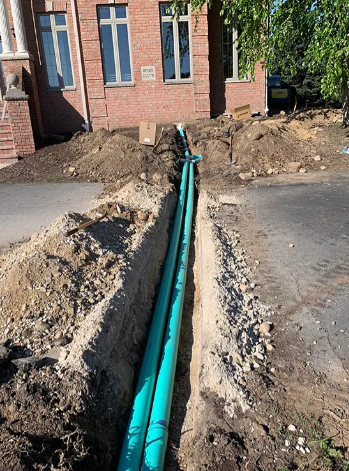 Two teal pipes laid inside a narrow dirt trench in front of a brick building.
