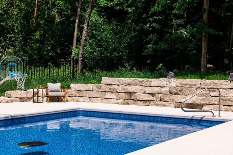 Swimming pool with stone retaining wall, chair, and basketball hoop in a wooded backyard.