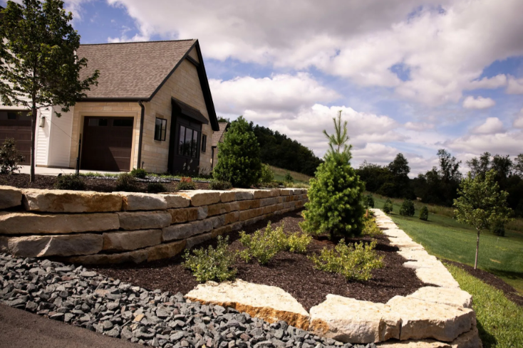 A stone retaining wall borders a garden bed with a small evergreen tree in front of a house under a cloudy blue sky.