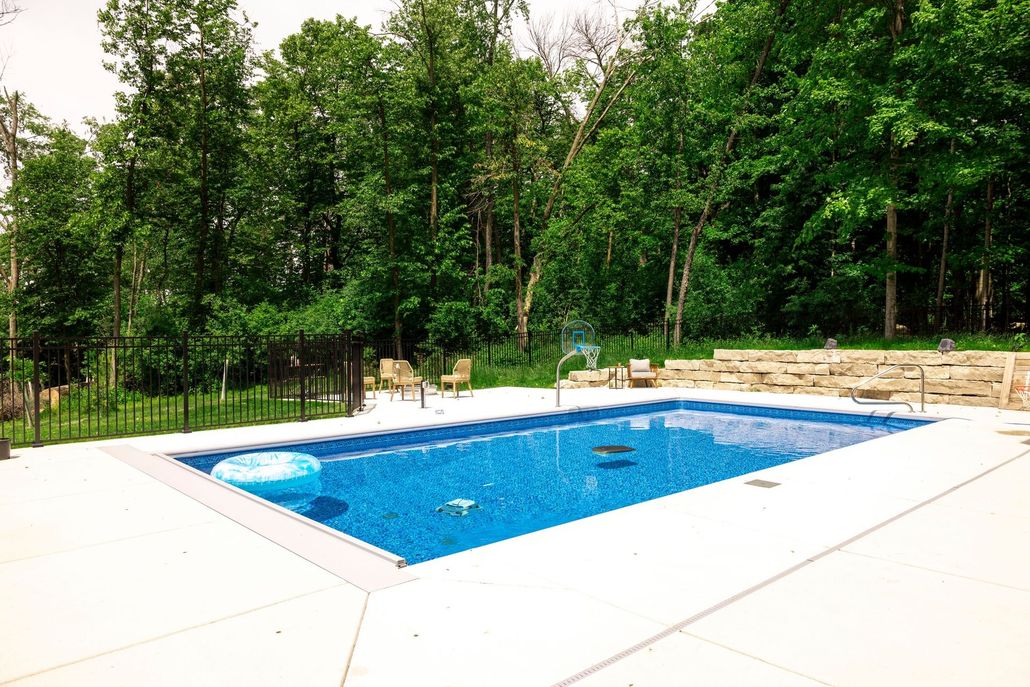 A rectangular blue swimming pool set in a backyard patio surrounded by dense green trees and a stone retaining wall.