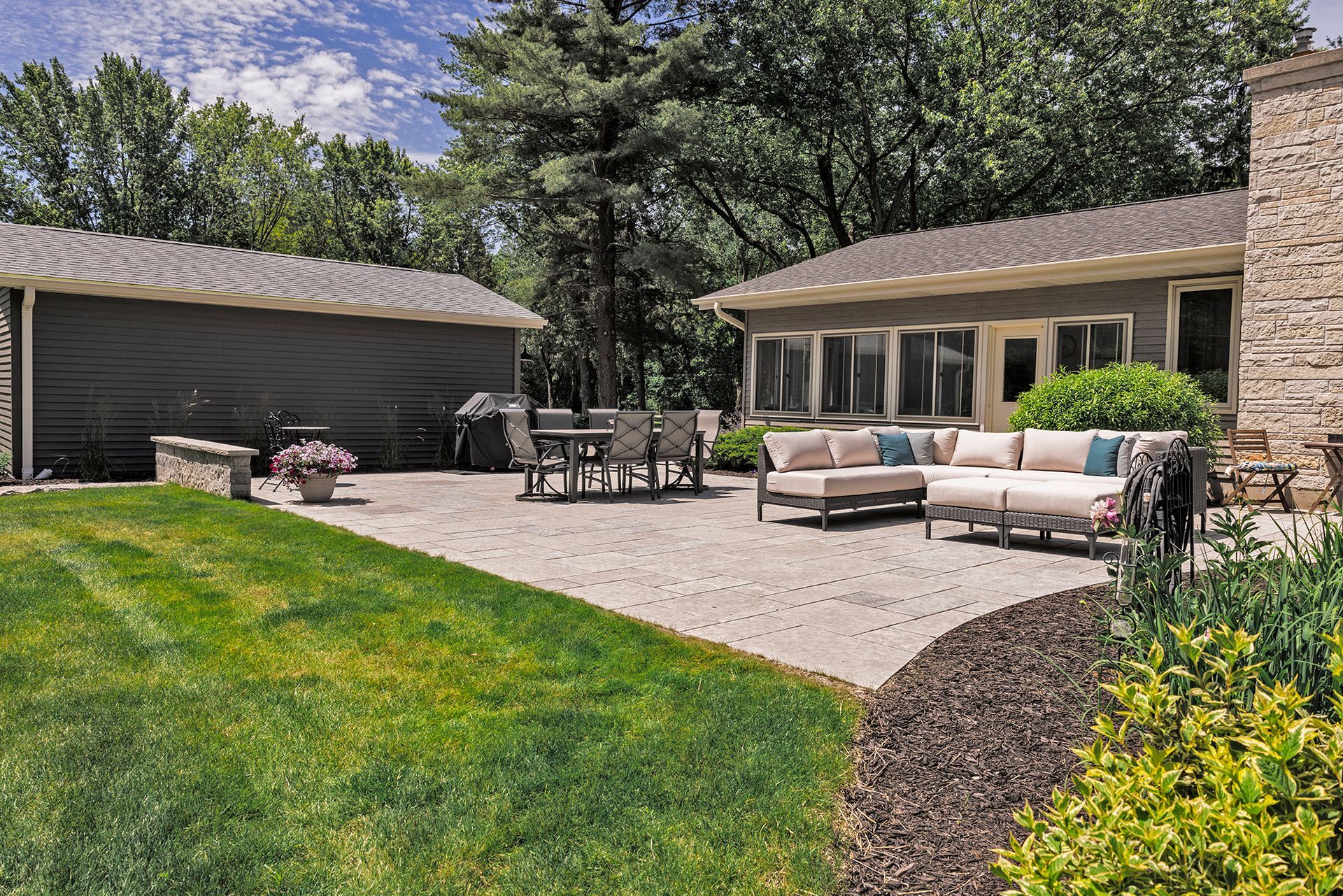 A sunny backyard patio features light-colored outdoor sectional seating, a dining table, and stone walls by a gray house.