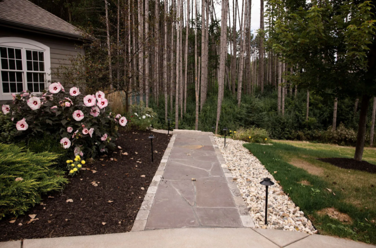 A stone walkway flanked by dark mulch with blooming flowers on the left and white decorative rocks on the right.