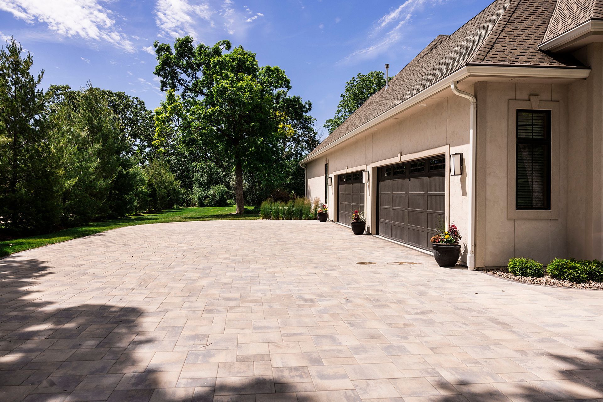 A beige house with a three-car garage and a large, paved stone driveway bordered by lush green trees under a blue sky.