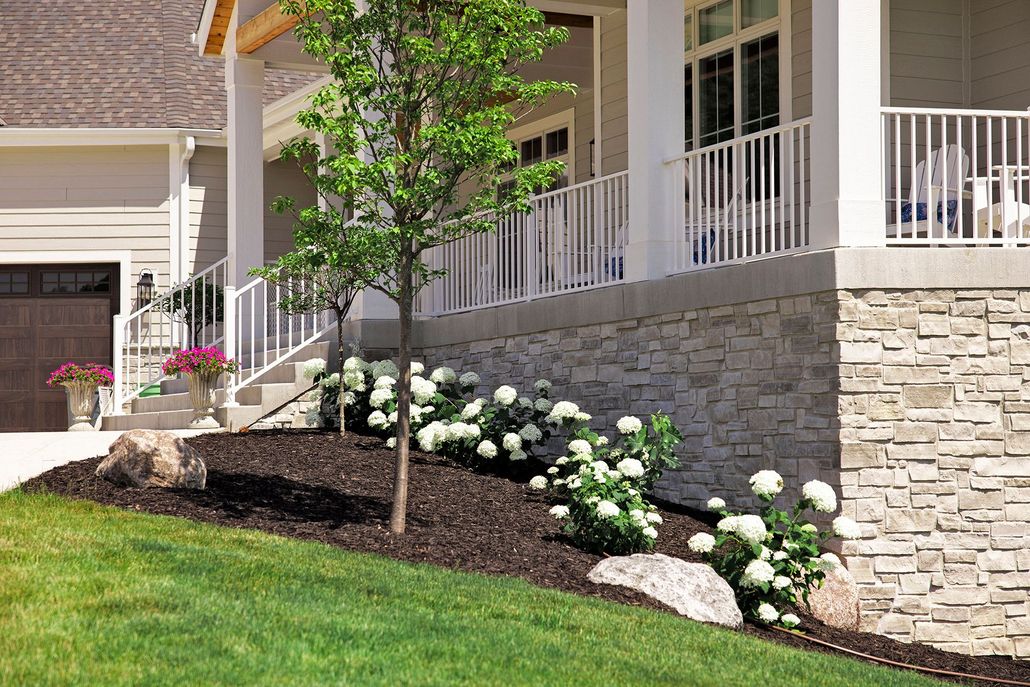 A stone-based porch with white railings, featuring a landscaped garden bed with mulch, white shrubs, and a young tree.
