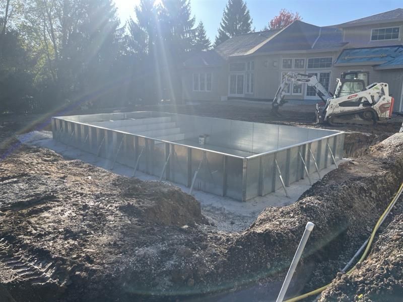 A rectangular metal swimming pool frame under construction in a residential backyard with a white skid steer nearby.