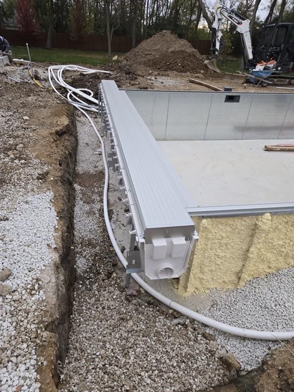 An in-ground pool under construction with metal walls, foam insulation, and plumbing lines visible in a dirt backyard.