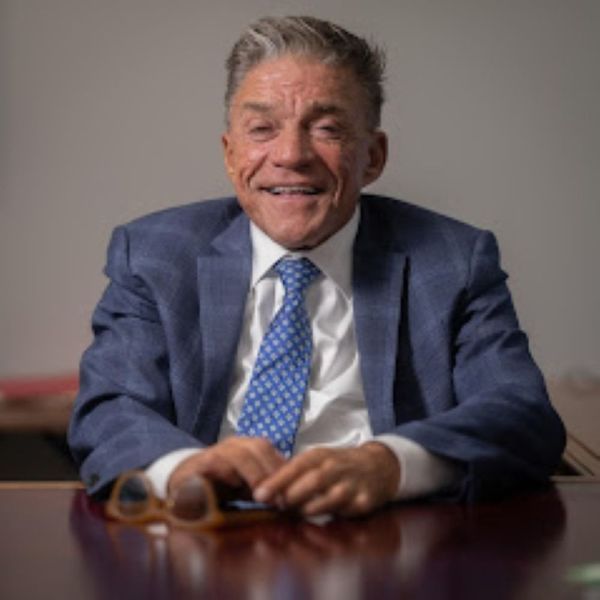 Man in blue suit, smiling, seated at a desk, holding glasses.