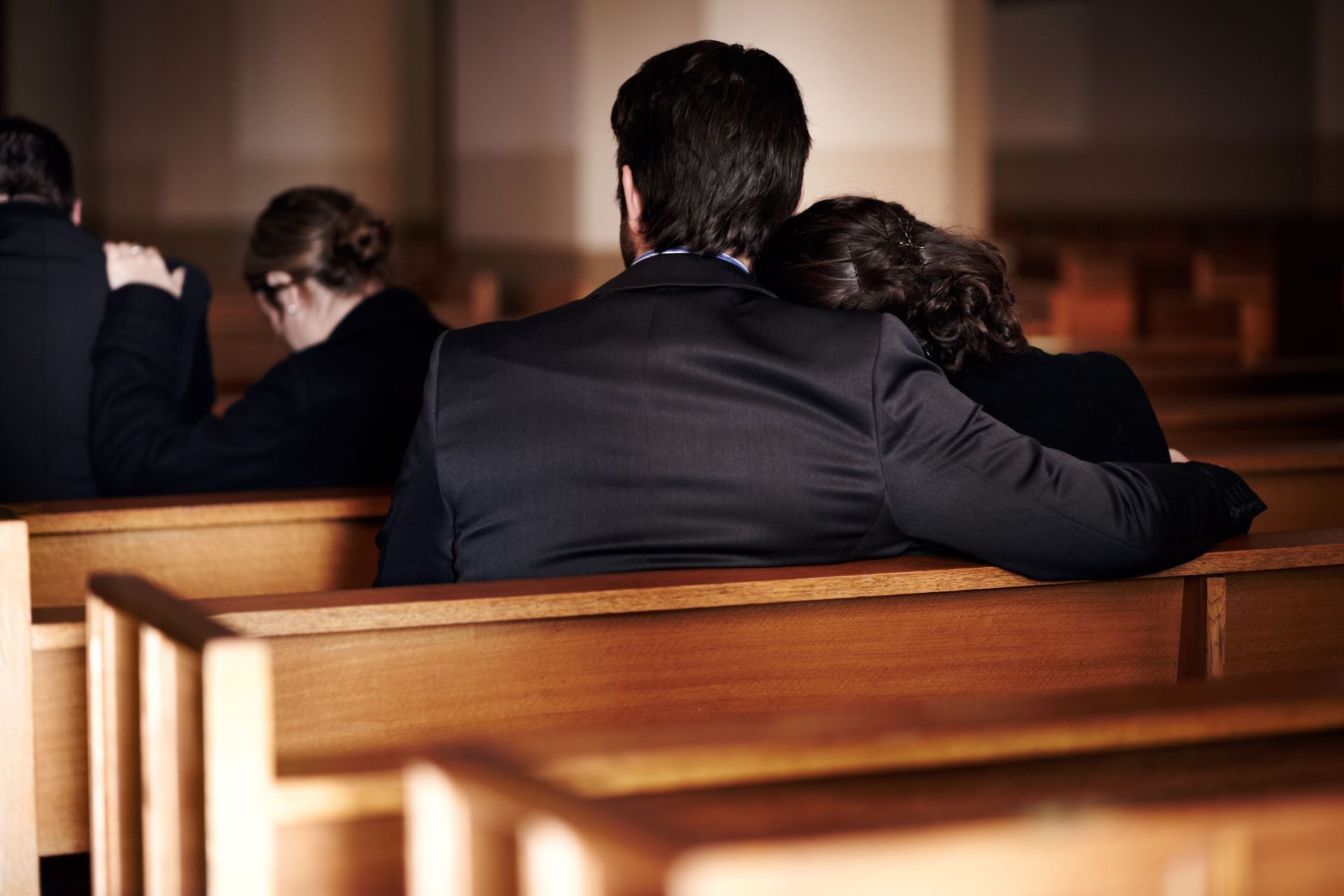 People in black attire sit in wooden pews, a woman leans on a man's shoulder. Church interior.