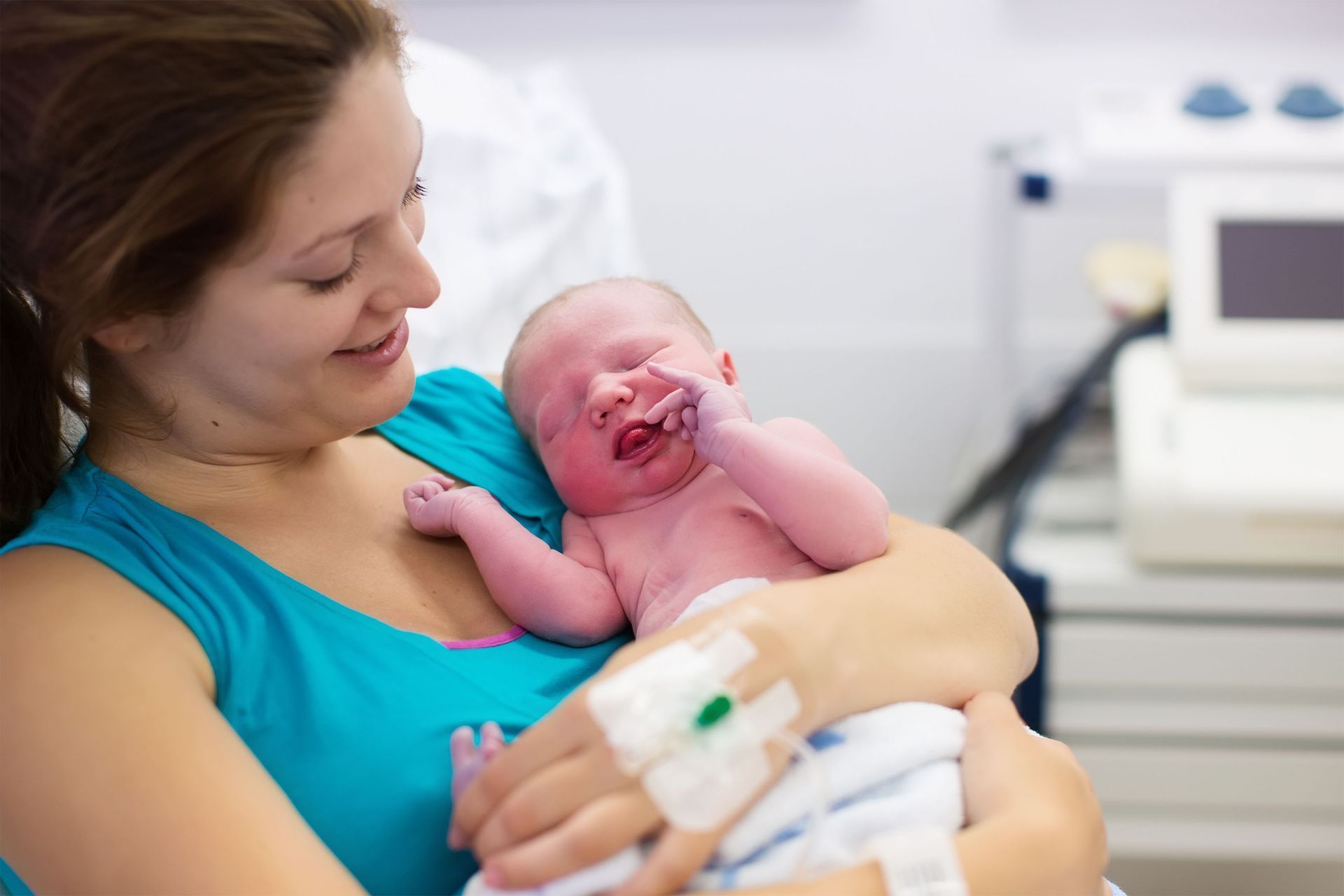 Woman holding a newborn baby in a hospital setting; baby's eyes closed, woman smiles.