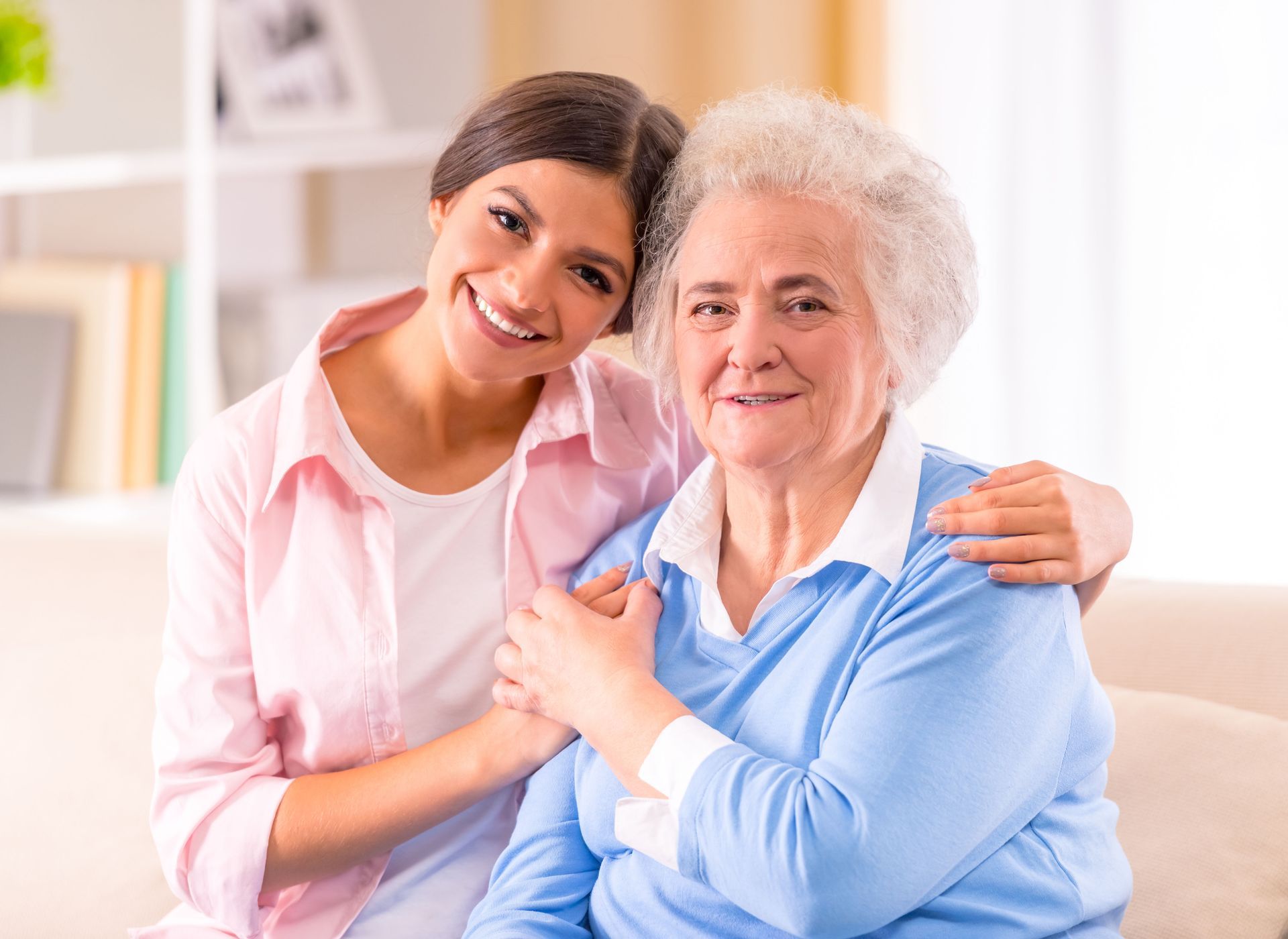 Woman hugs an older person, both smiling; indoor setting with neutral background.