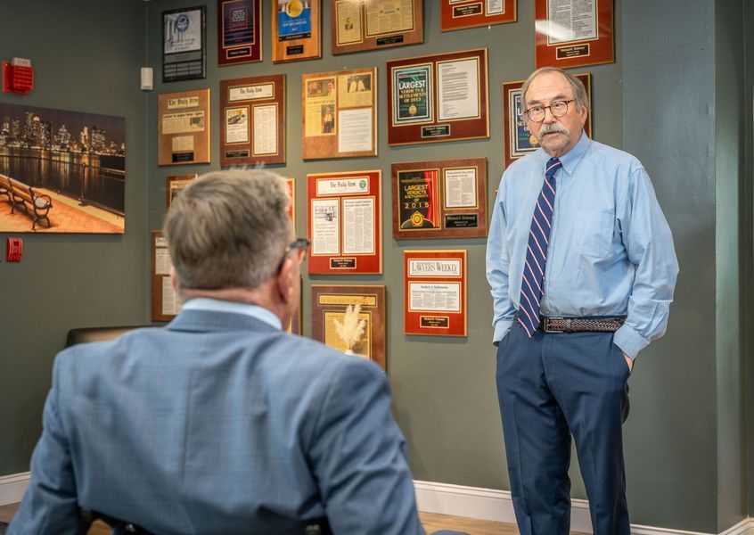 Man in blue shirt, hands in pockets, stands near awards wall, facing person in blue suit seated.