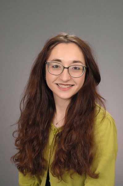 Woman wearing glasses smiles at the camera, with long, wavy brown hair, wearing a yellow cardigan.