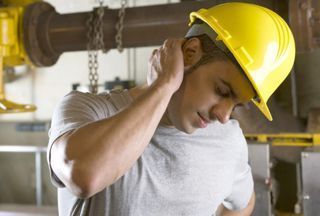 Man in yellow hard hat holding his neck in apparent pain at a worksite.