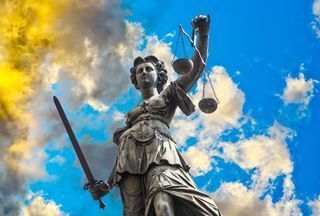 Statue of Lady Justice holding scales and a sword against a cloudy blue sky.