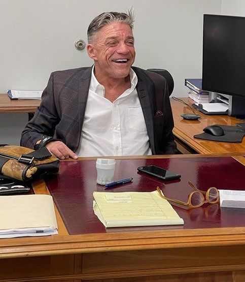 Man smiling at a desk, wearing a jacket and white shirt. Office setting with documents and a computer.