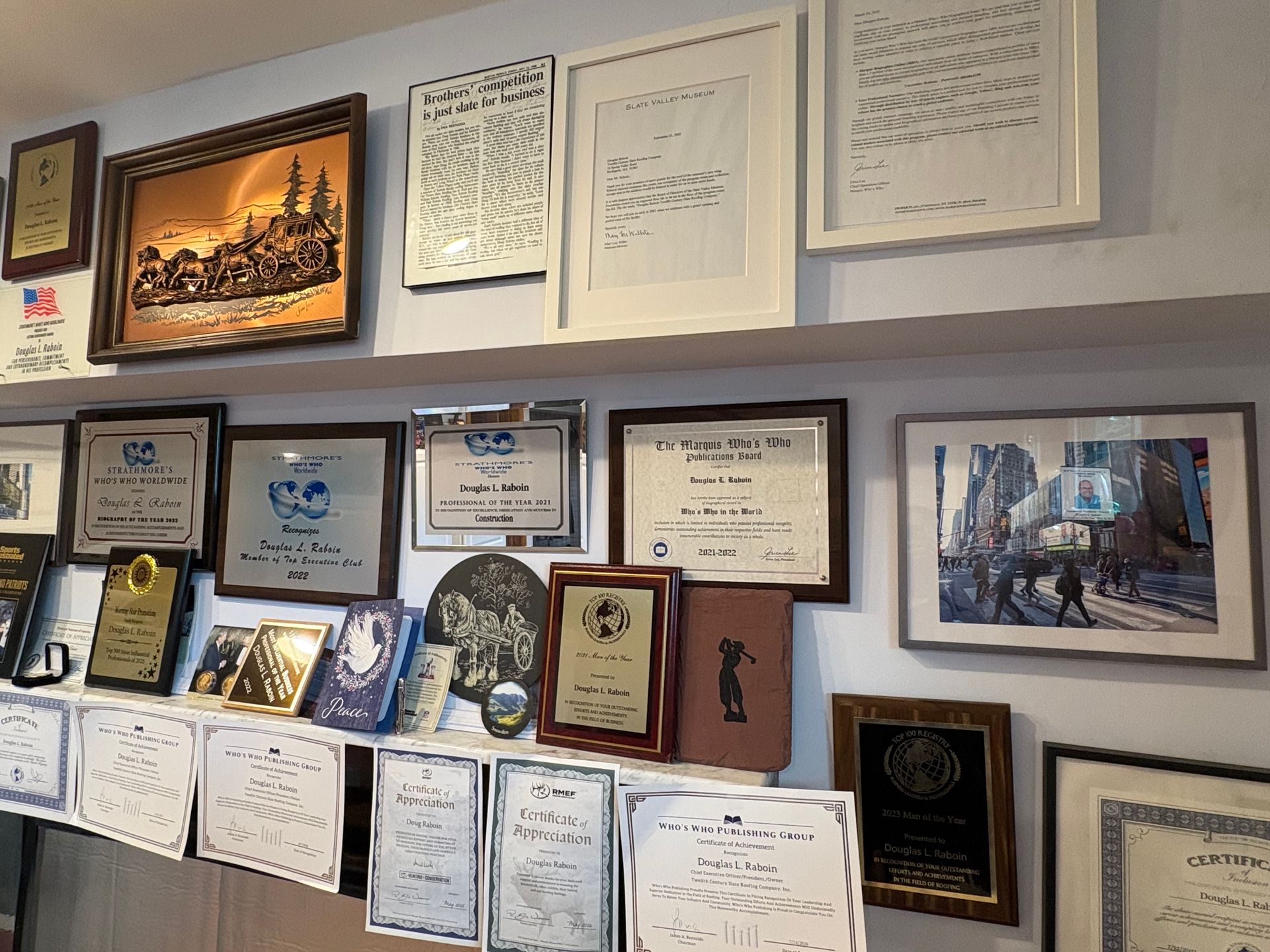 Wall of framed certificates, awards, and artwork on a light-colored office wall.