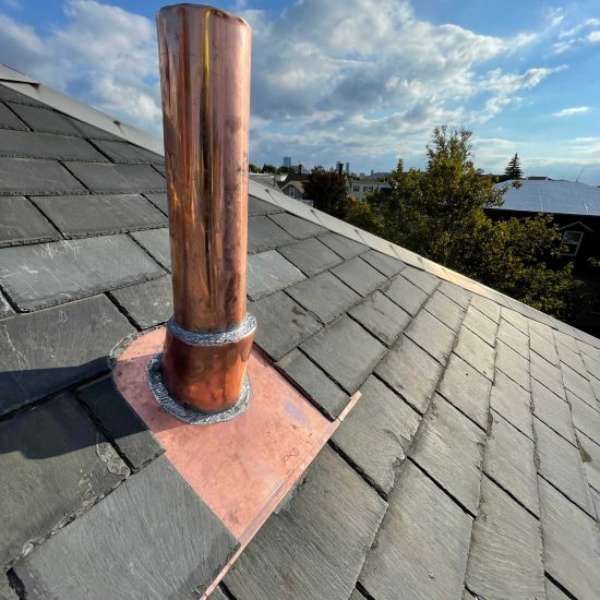 Copper chimney pipe on a gray shingled roof under a partly cloudy sky.