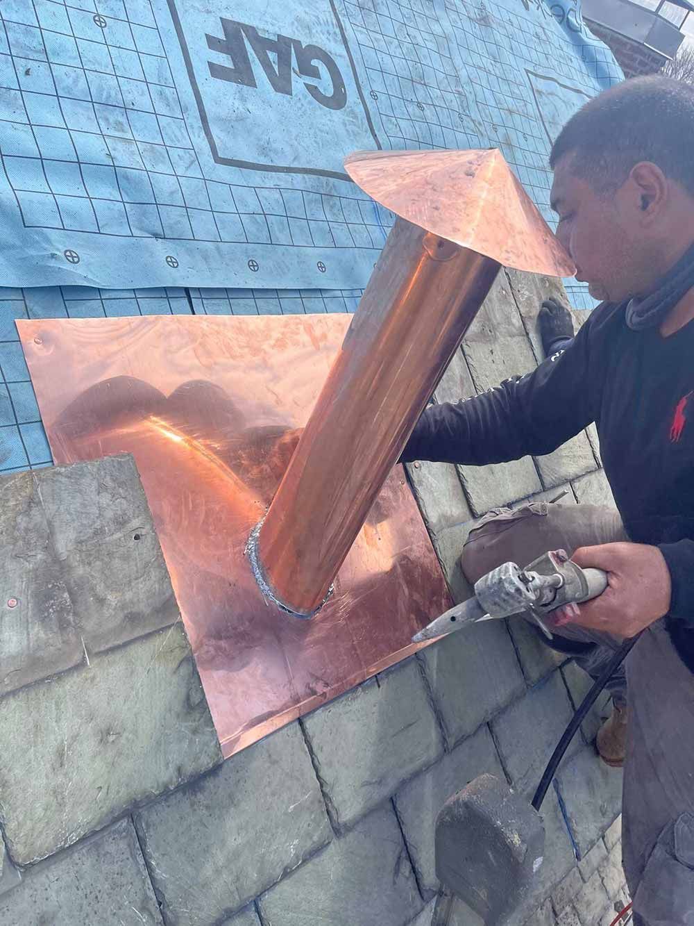 Worker installing a copper roof vent flashing on a rooftop with a torch.