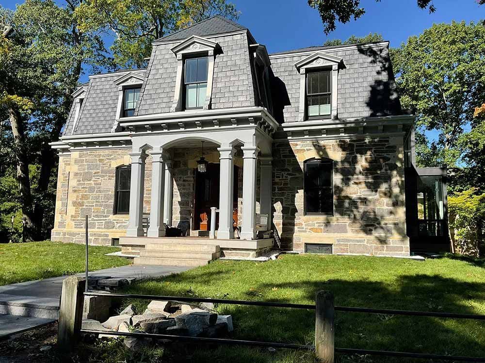 Stone house with a gray mansard roof and front columns, set on a sunny lawn with trees.