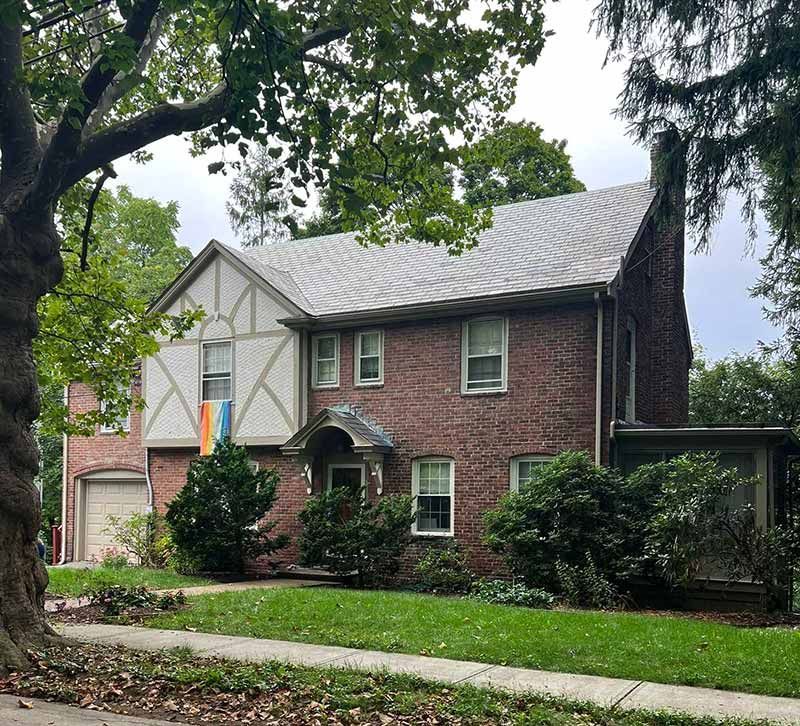 Two-story brick and stucco house with a front lawn, trees, and an attached garage on a quiet street
