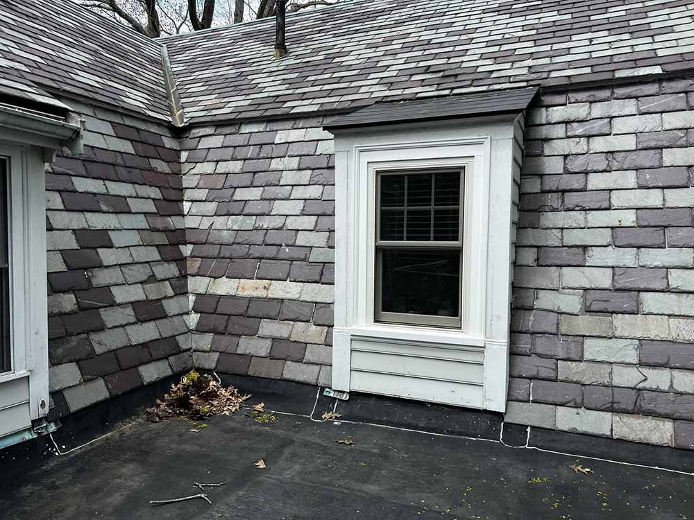 Gray shingled house exterior with a white-trimmed window and dark roof at a corner of the building