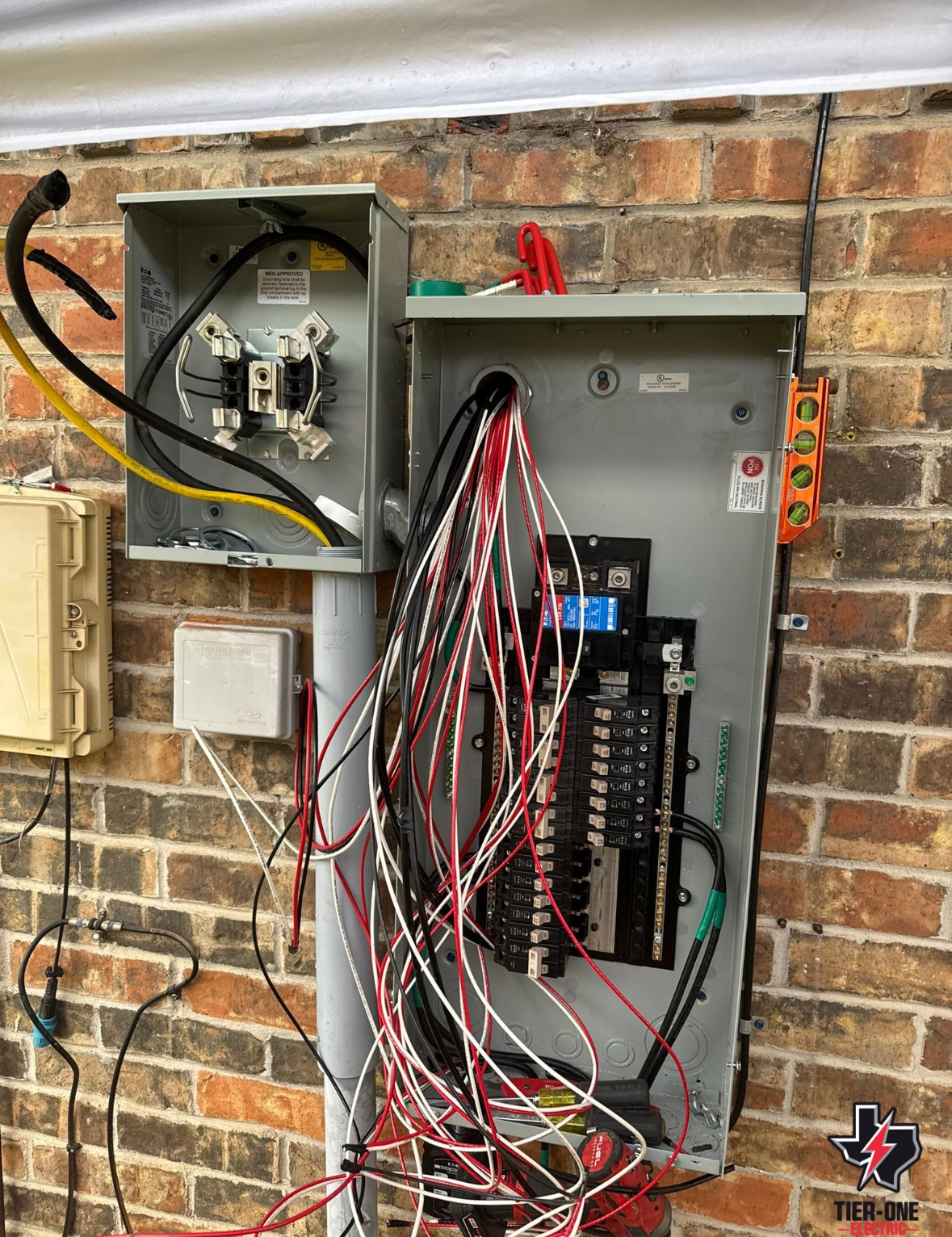 Electrical service panel with wires exposed, mounted on a brick wall, next to a utility box.