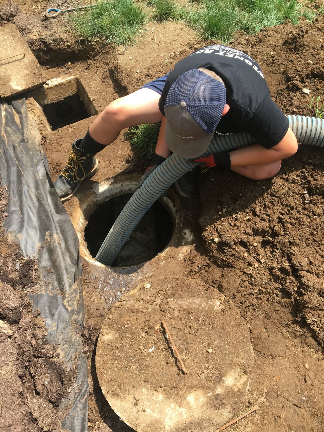 A man is kneeling down in the dirt and pumping water into a septic tank.