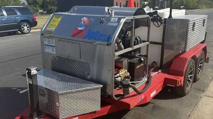Trailer-mounted industrial power washer; red frame, silver machinery, black tires, parked on asphalt.