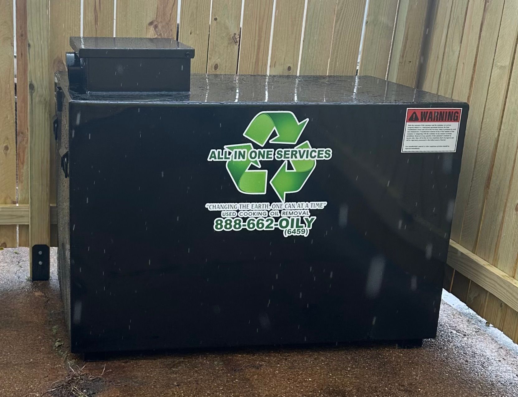 Black recycling bin with green logo and warning labels, set against a wooden fence.