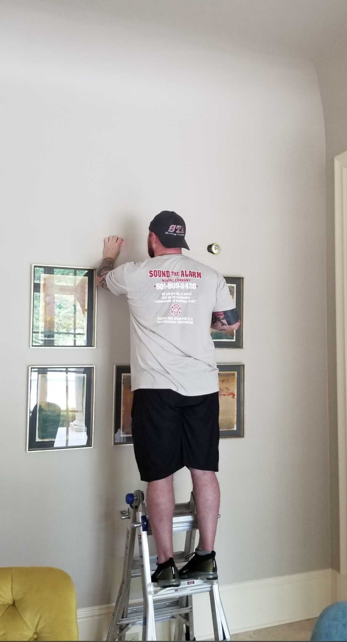 Person on a stepladder hangs framed pictures on a light gray wall.