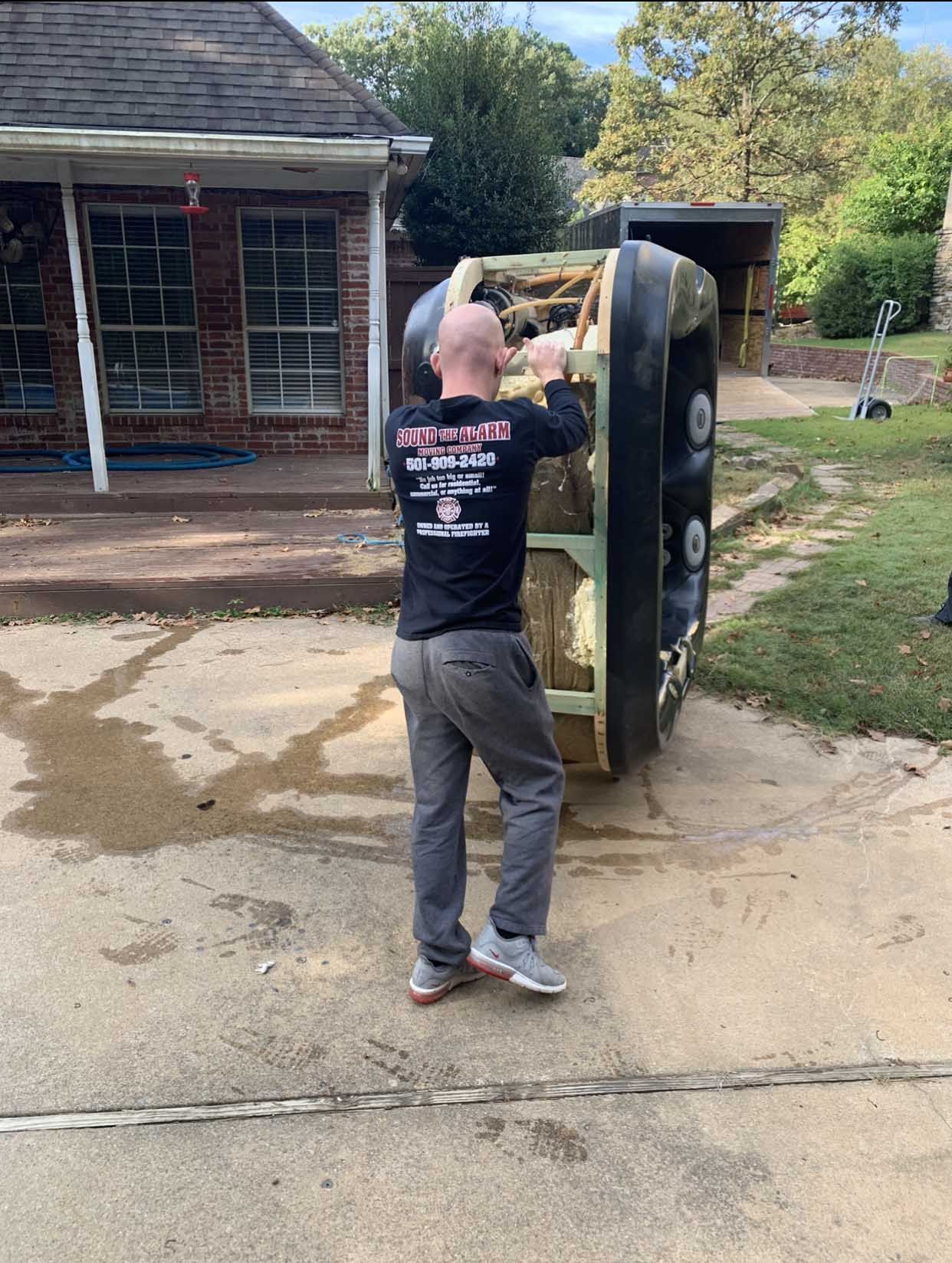 Man lifting a large black dumpster outside a brick building.