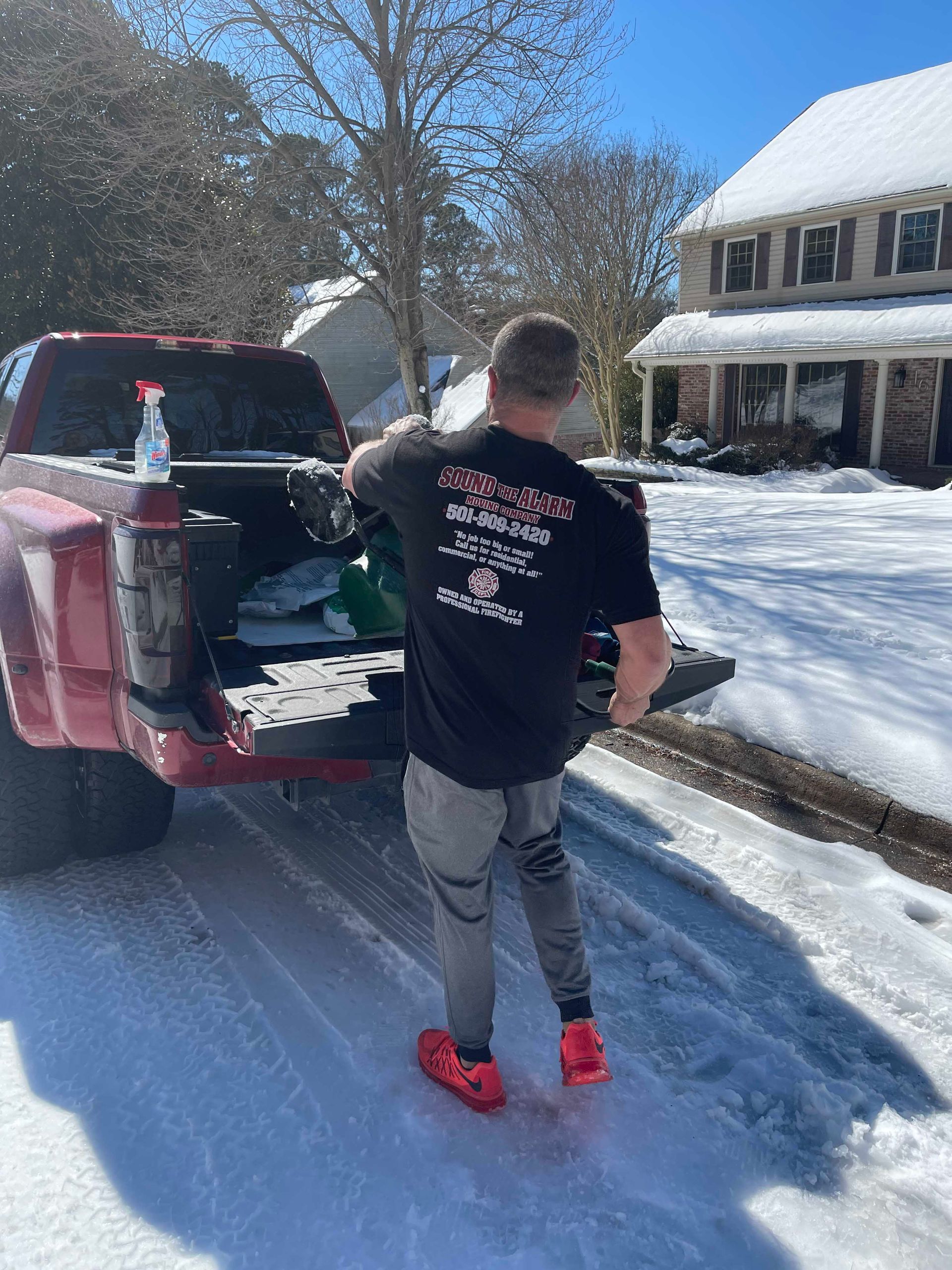 Man in black shirt with logo unloading truck bed in snowy residential area.