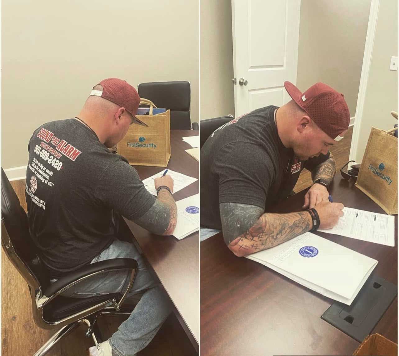 Man in a cap, tattoos, signing documents at a desk in an office.