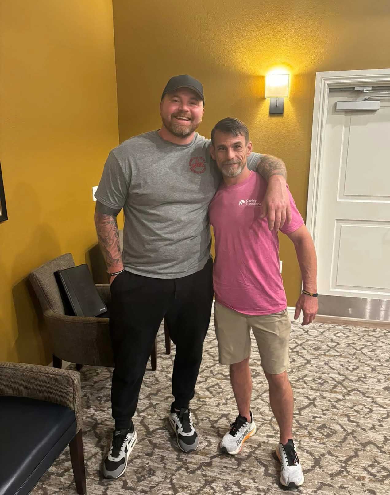 Two men pose for a photo. One tall man in gray shirt and black pants, another in pink shirt and khaki shorts.