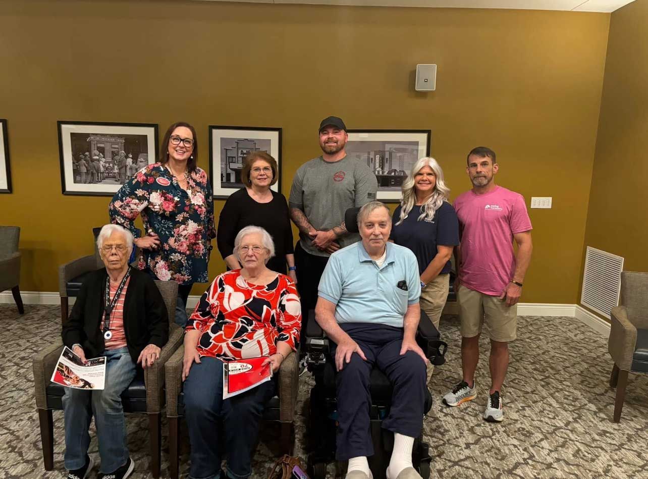 Group of people posing for a photo indoors. Some seated, some standing, in front of a wall with artwork.