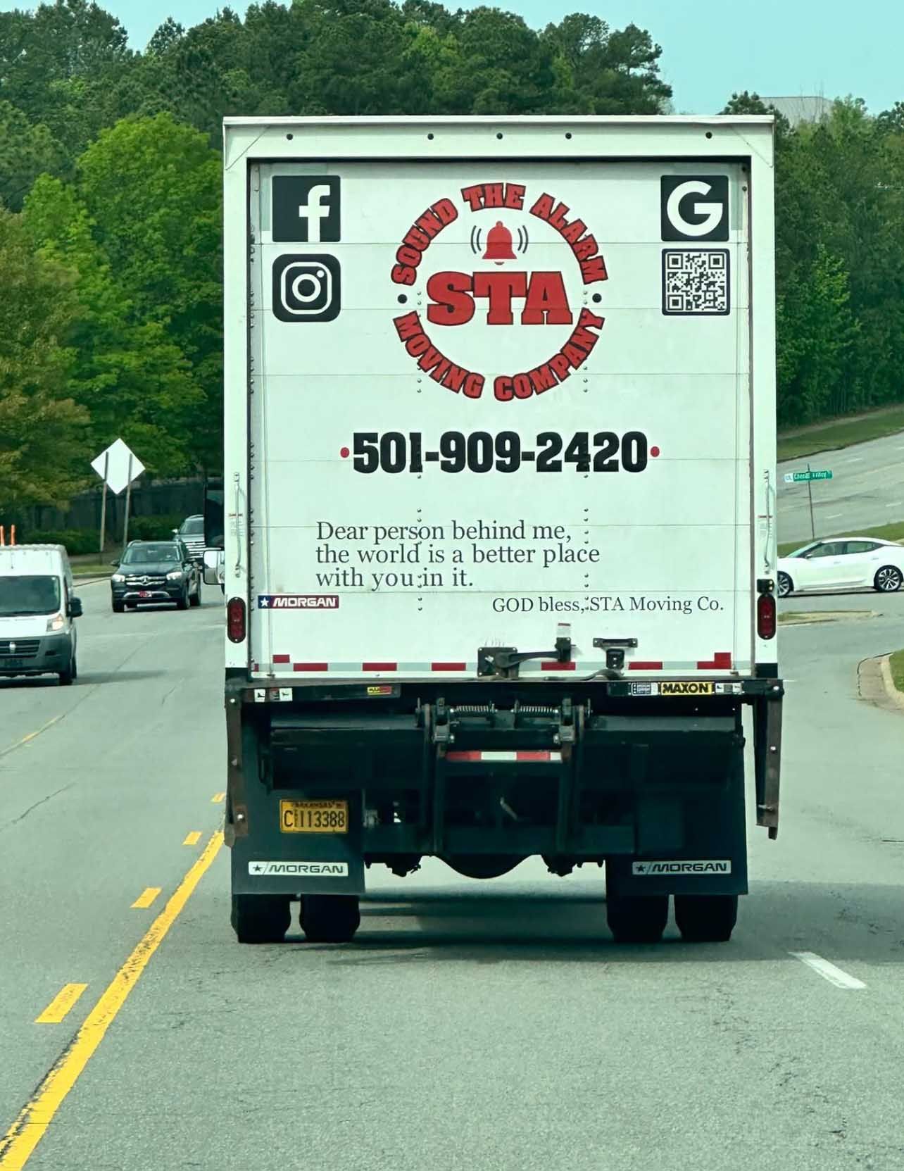 Back of a white moving truck with logo and contact info on a road, other vehicles in background.