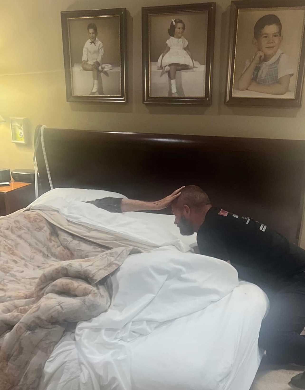 Man kneeling at bedside, hand on arm on bed. Three framed portraits hang above the bed.