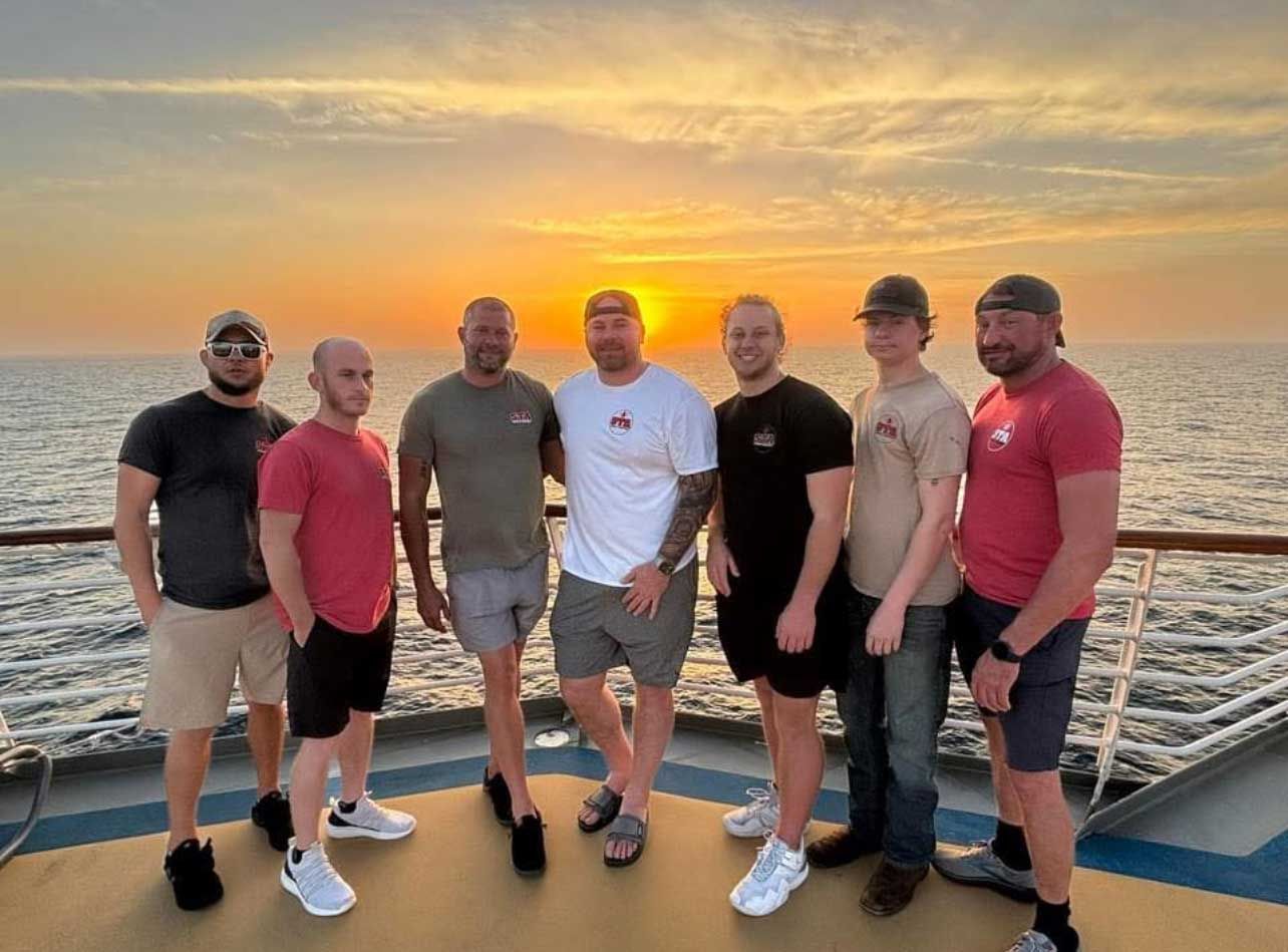 Group of men on a ship deck at sunset. Some wear t-shirts and shorts. Golden sky over ocean.