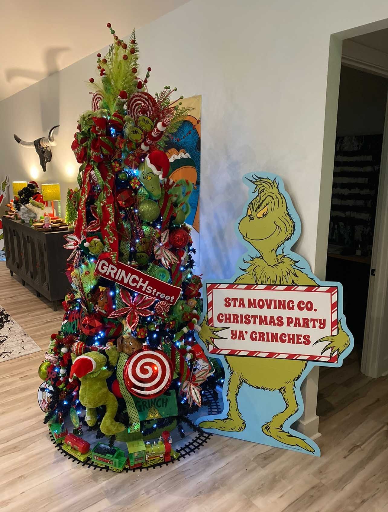 Christmas tree decorated with Grinch-themed ornaments next to a Grinch cutout advertising a Christmas party.