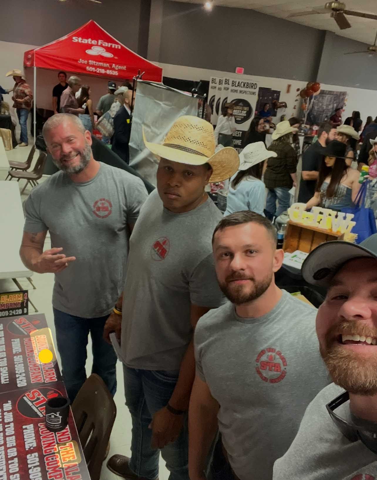 Four men wearing matching grey shirts and cowboy hats smile at the camera at a fair.