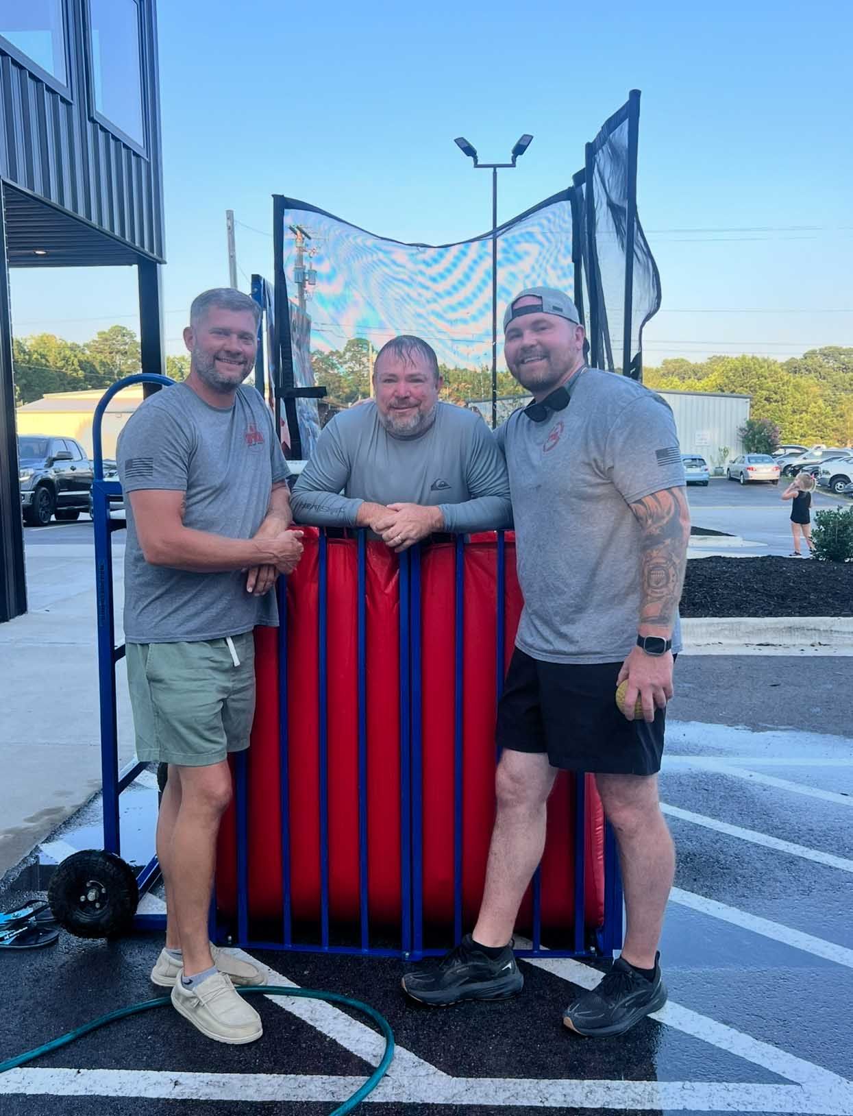 Three men stand by a red, blue, and black dunk tank outdoors. Two wear shorts and gray shirts.
