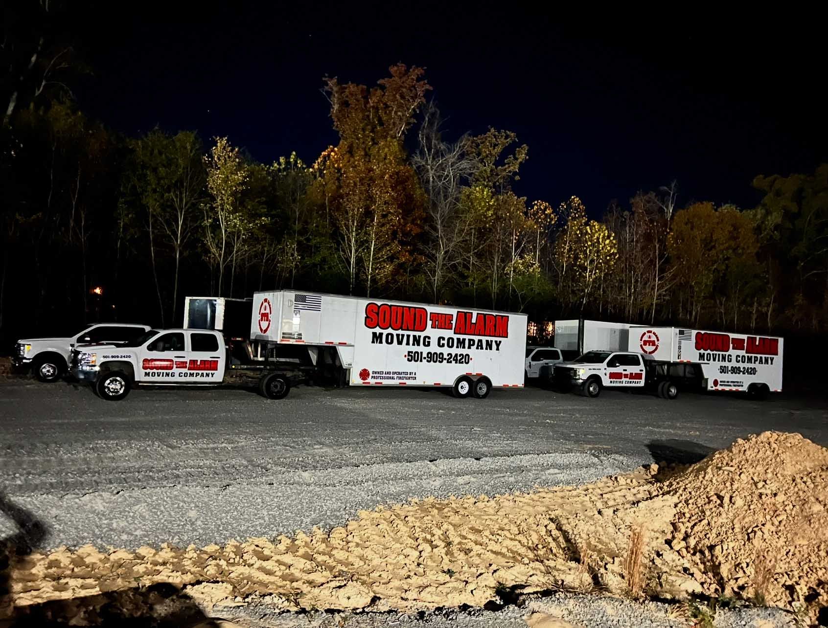 Moving trucks and service vehicles parked in gravel lot at night. Trees in background.